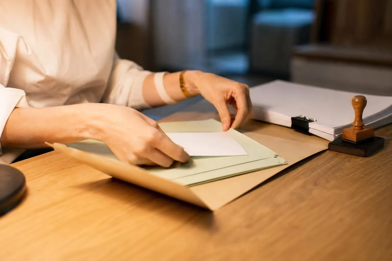 Les mains d’une assistante juridique glissent une feuille entre deux sous-chemises dans une chemise à rabats ouverte sur une table de chêne clair.